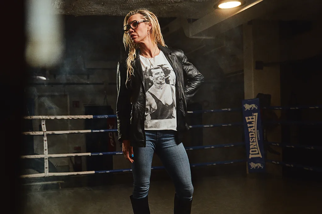 Mikaela Lauren med glasögon, svart skinnjacka, jeans och stövlar står i en boxningsring. På hennes T-shirt syns ett tryck av en boxare. Bakgrunden är svagt upplyst, med rök och en boxningsring av märket Lonsdale synlig.