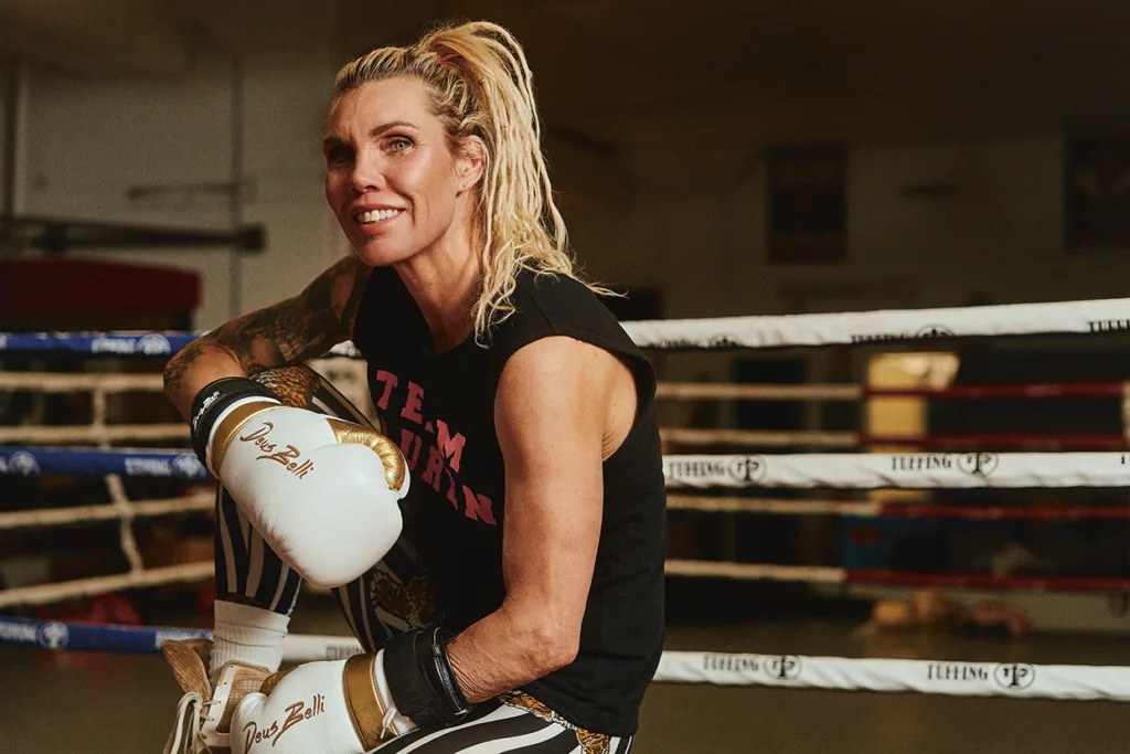 En leende Mikaela Laurén med blont hår i hästsvans, iklädd boxningshandskar, en ärmlös Team-tröja och randiga leggings, sitter på kanten av en boxningsring inne på ett gym.