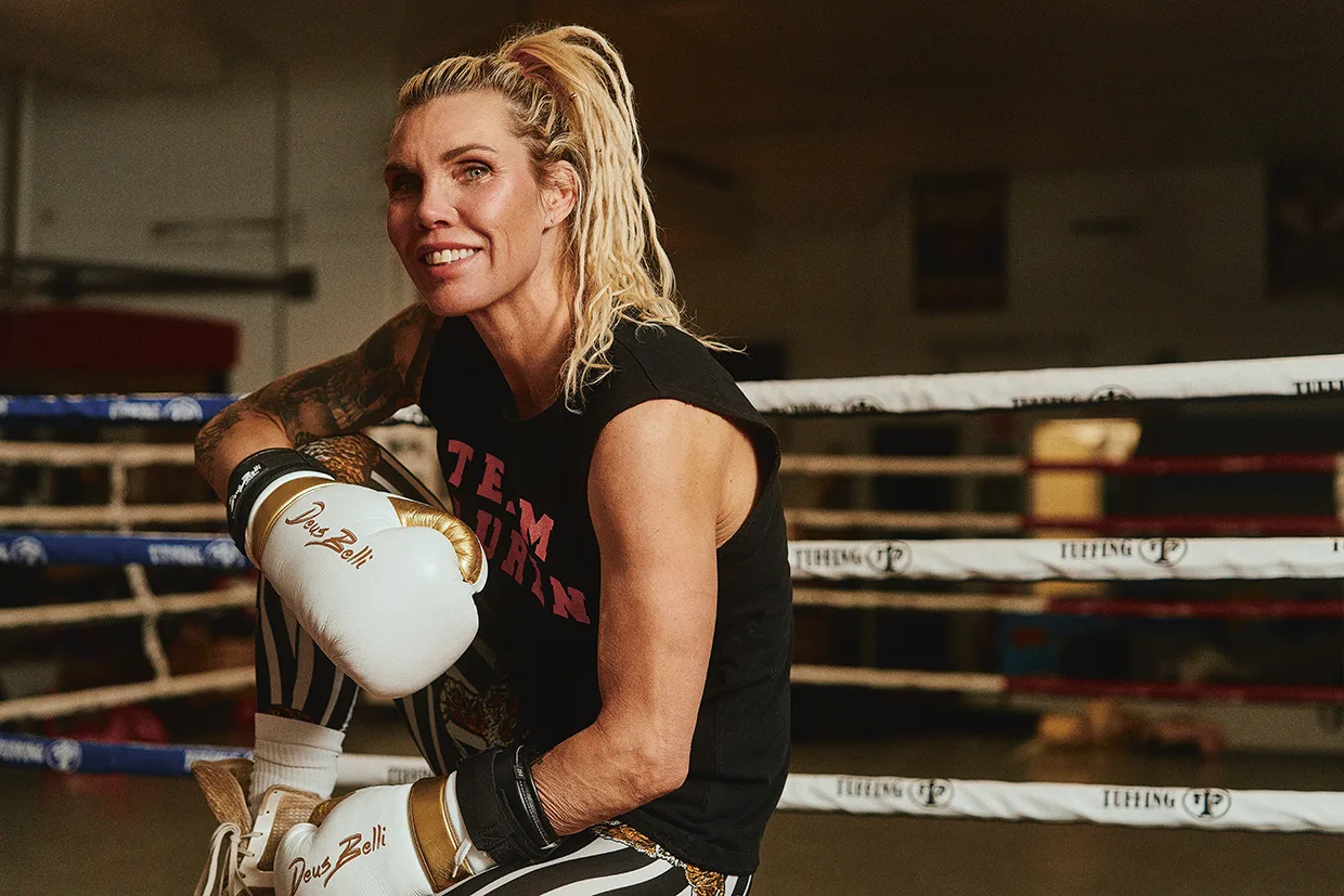 En leende Mikaela Laurén med blont hår i hästsvans, iklädd boxningshandskar, en ärmlös Team-tröja och randiga leggings, sitter på kanten av en boxningsring inne på ett gym.