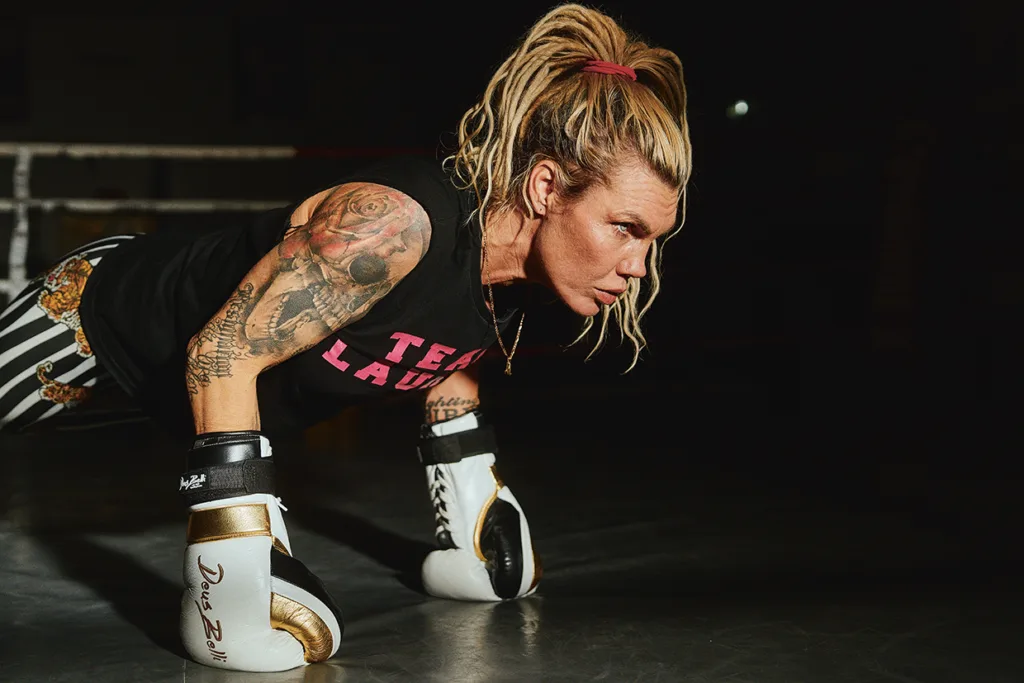 En fokuserad Mikaela Lauren med tatueringar och boxningshandskar gör armhävningar i ett svagt upplyst boxningsgym, klädd i en svart TEAM LAURÉN-tröja och randiga shorts, med håret uppsatt i en hög hästsvans.