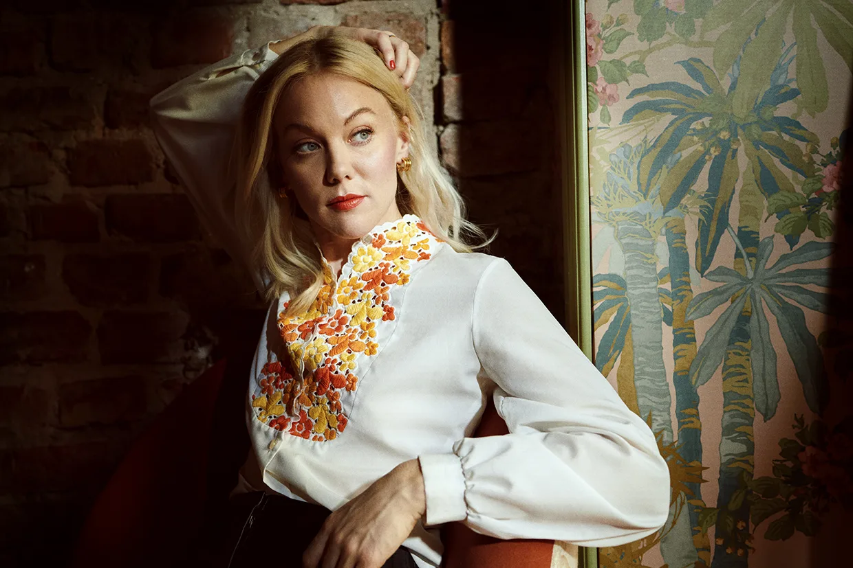 Agnes Lindström Bolmgren med blont hår och vit blus med orange och gula blommor sitter i en stol och vilar armen på huvudet. Hon tittar åt sidan. Bakom henne finns en tegelvägg och en färgglad tapet med bladverk.