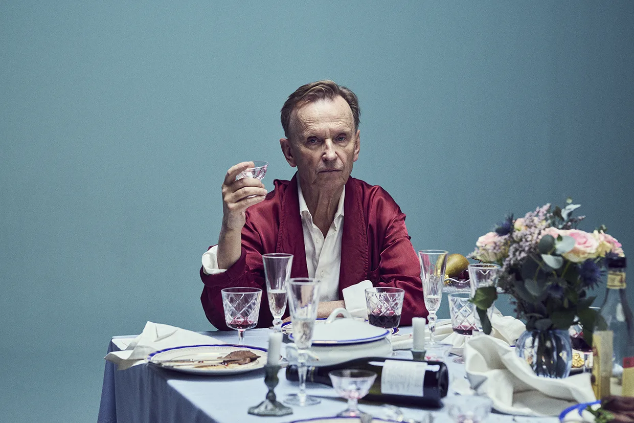 Johan Ulveson i röd mantel sitter ensam vid ett stökigt, elegant dukat matbord och håller i ett glas. Bordet är täckt av tomma glas, tallrikar, en vinflaska och en blommig mittpunkt mot en blå bakgrund.