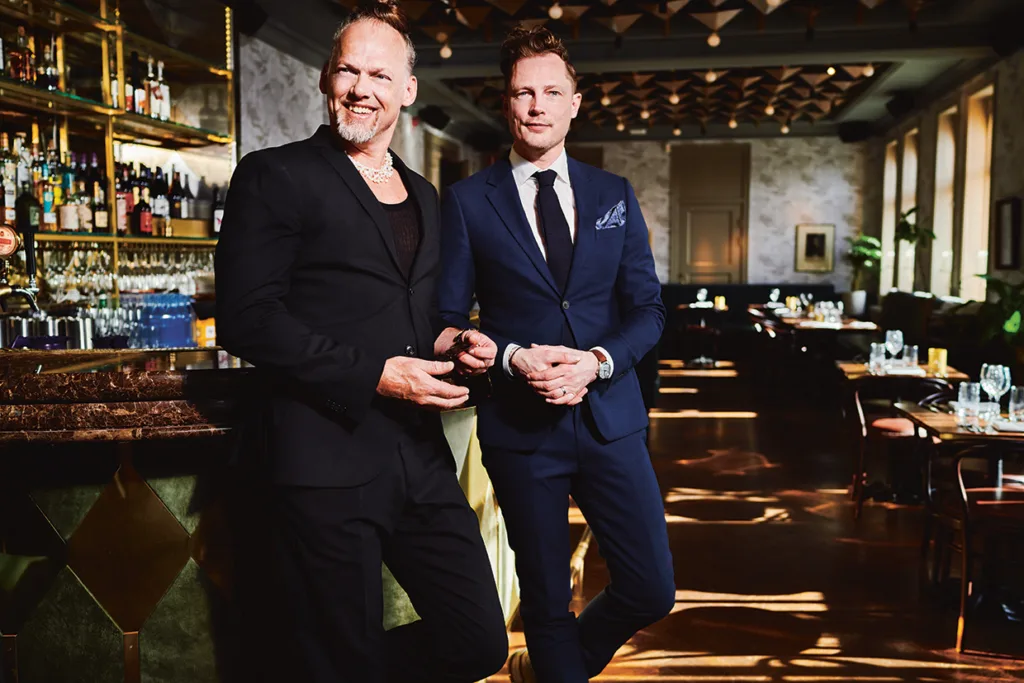Kostymklädda Brynolf & Ljung står självsäkra i en stilfull, exklusiv restaurang med elegant inredning, en välfylld bar och varm belysning. Den ene mannen lutar sig mot baren medan den andre står bredvid honom, båda ler.