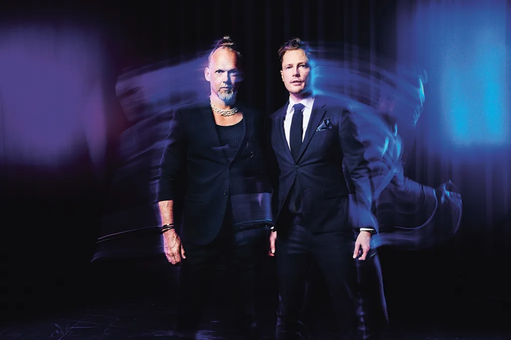 Brynolf & Ljung i formella kostymer står sida vid sida mot en mörk, suddig bakgrund med lila och blå belysning. Den ene mannen har skägg och en toppknut, den andre är slätrakad och har kort hår. De omges av rörelseoskärpa.