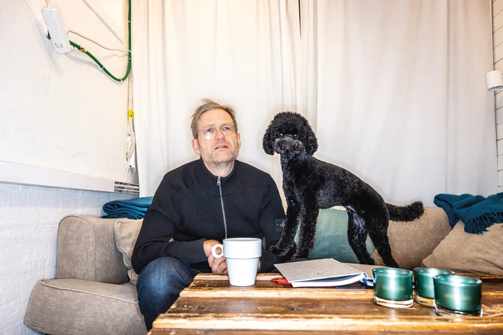 En man sitter i en soffa med en mugg i handen och tittar eftertänksamt upp. En svart pudel står bredvid honom på ett soffbord i trä, där det ligger en anteckningsbok, en penna och ljus. Bakom dem hänger en vit gardin.