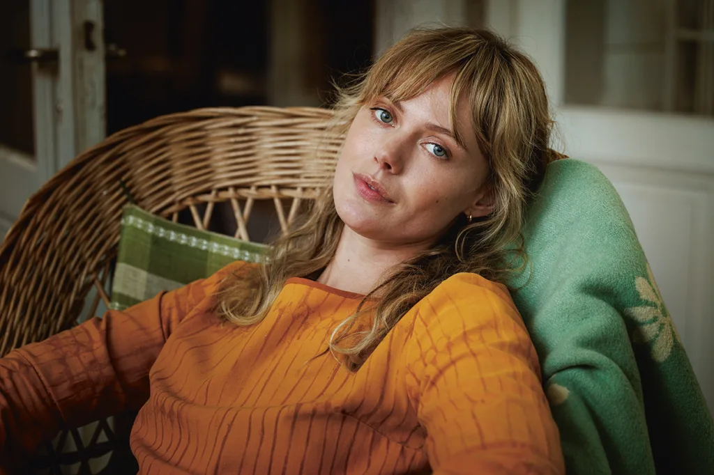 Frida Gustavsson med blont hår och blå ögon, klädd i en orange topp, sitter i en korgstol, lutar sig tillbaka mot en grön filt och tittar eftertänksamt in i kameran.