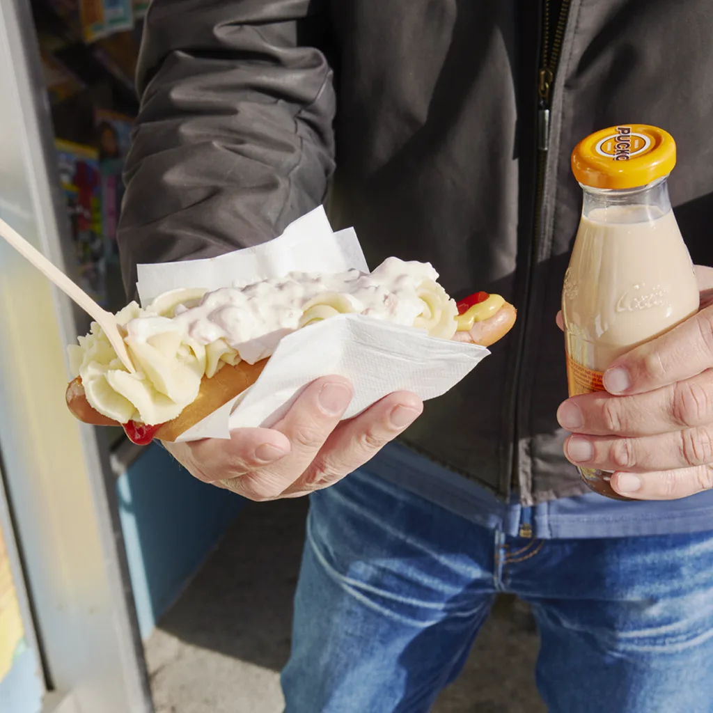 En person i mörk jacka och jeans står utomhus och håller en varmkorv med lök och sås i ena handen och en flaska chokladmjölk i den andra.