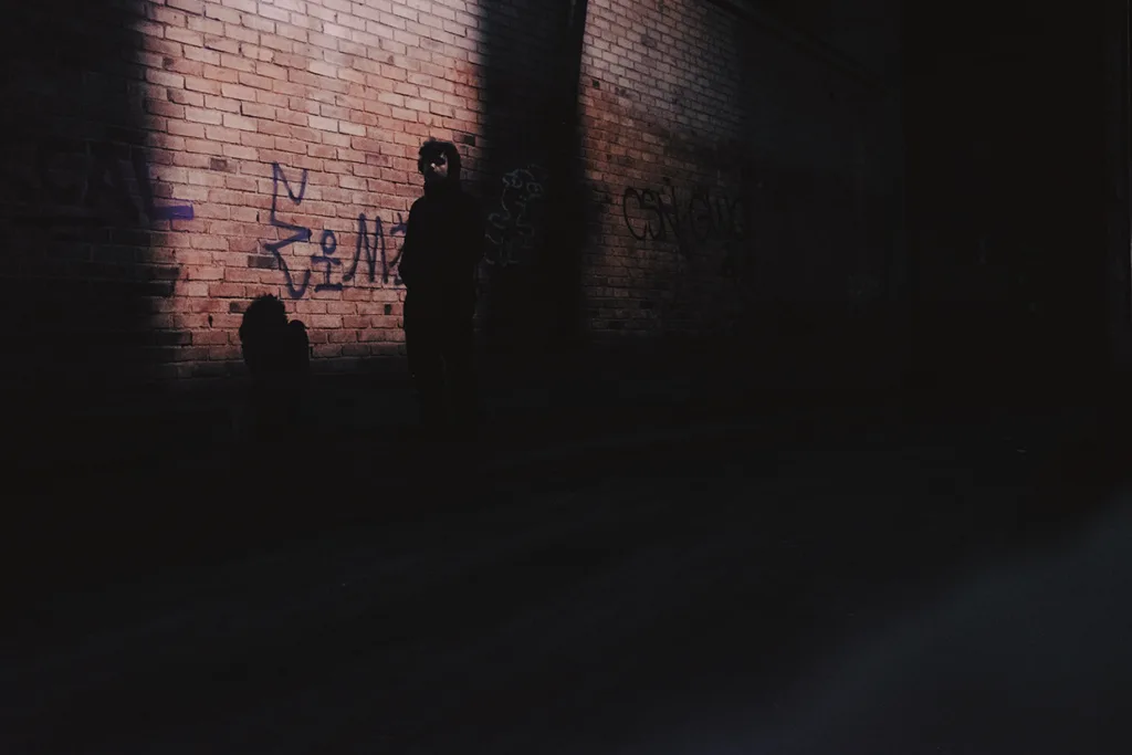 En person står i en skuggig gränd mot en tegelvägg med graffiti, delvis upplyst av en ljusstråle medan större delen av scenen förblir mörk.