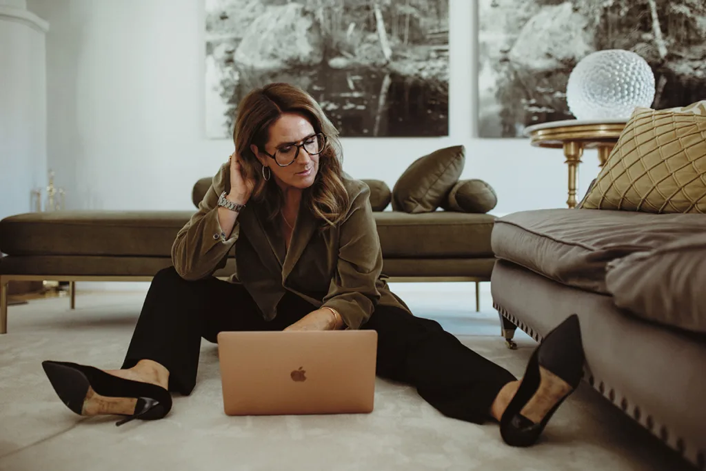 Denise Rudberg med långt brunt hår och glasögon sitter på golvet mellan en soffa och ett soffbord och arbetar på en laptop. Hon bär en grön kavaj, svarta byxor och svarta klackar, med modern inredning i bakgrunden.