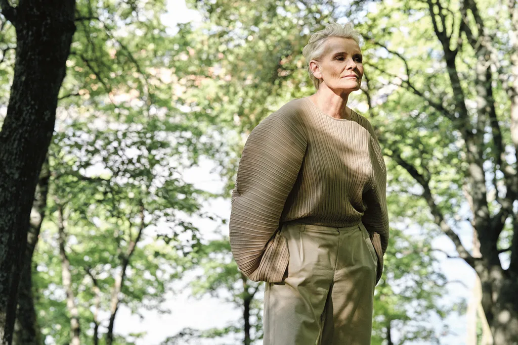 En äldre person med kort blont hår står i en solbelyst skog, klädd i en lös, ribbstickad beige topp med vida ärmar och solbruna byxor med hög midja, händerna i fickorna, och tittar eftertänksamt ut i fjärran.