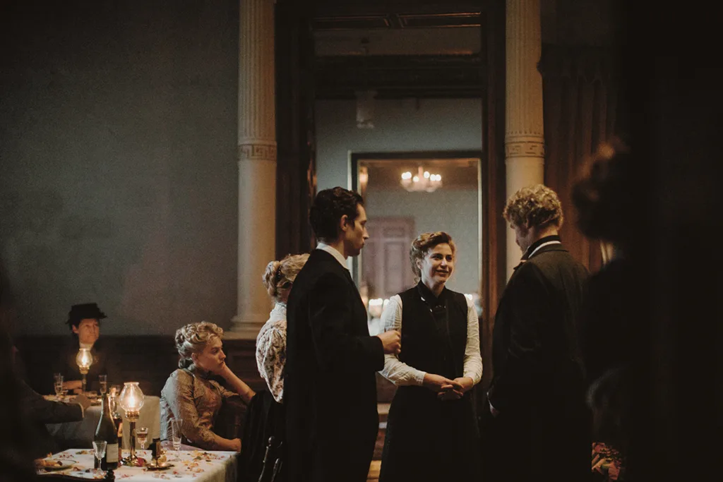 En grupp människor i vintagekläder står och samtalar i ett svagt upplyst, elegant rum med höga pelare, medan andra sitter vid ett bord dukat med flaskor och ljus.