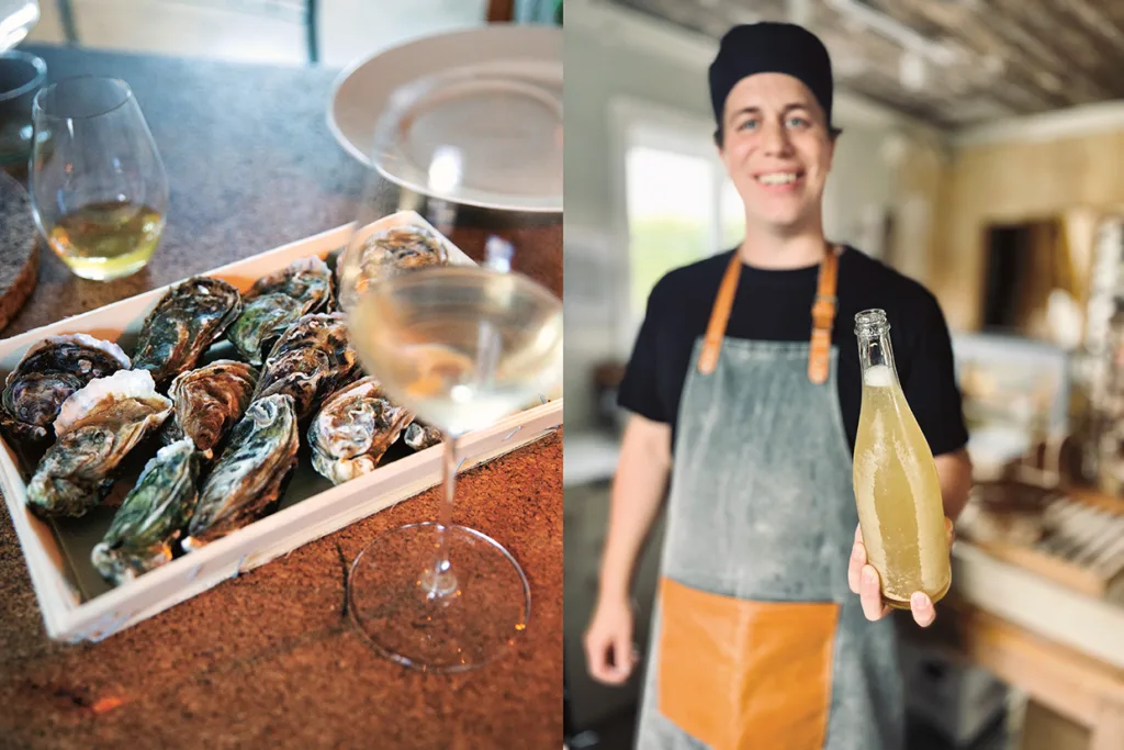 Närbild av en bricka med ostron och två glas vitt vin på ett bord, bredvid en leende person i förkläde och hatt som håller i en flaska med ljus dryck i ett ljust kök.