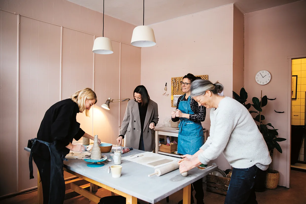 Fyra kvinnor samlas runt ett bord i ett mysigt, väl upplyst rum och arbetar med konst- eller hantverksprojekt med lera och verktyg, ler och njuter av varandras sällskap. Hyllor, en växt och en klocka dekorerar bakgrunden.
