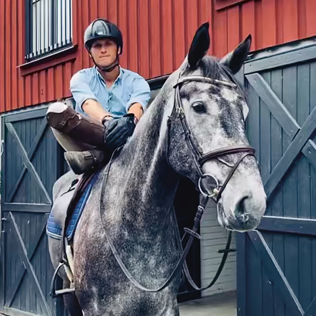 Carl Hedin med hjälm och blå skjorta sitter på en grå häst framför en röd byggnad med stora ladugårdsdörrar. Hästen har sadel och träns och ryttaren håller i tyglarna.