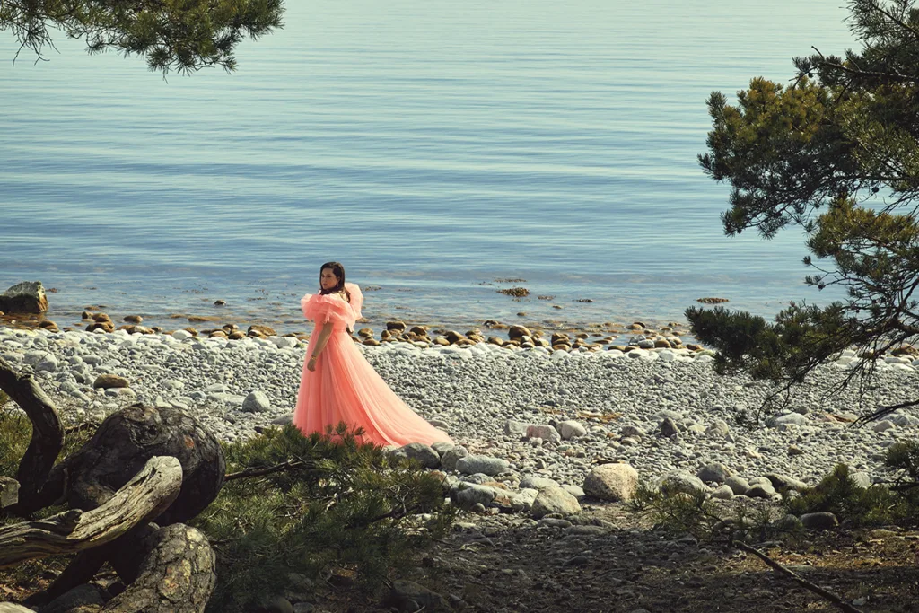 Viktoria Tocca i en rosa klänning står på en klippig strand nära lugnt vatten, inramad av trädgrenar i förgrunden en solig dag.