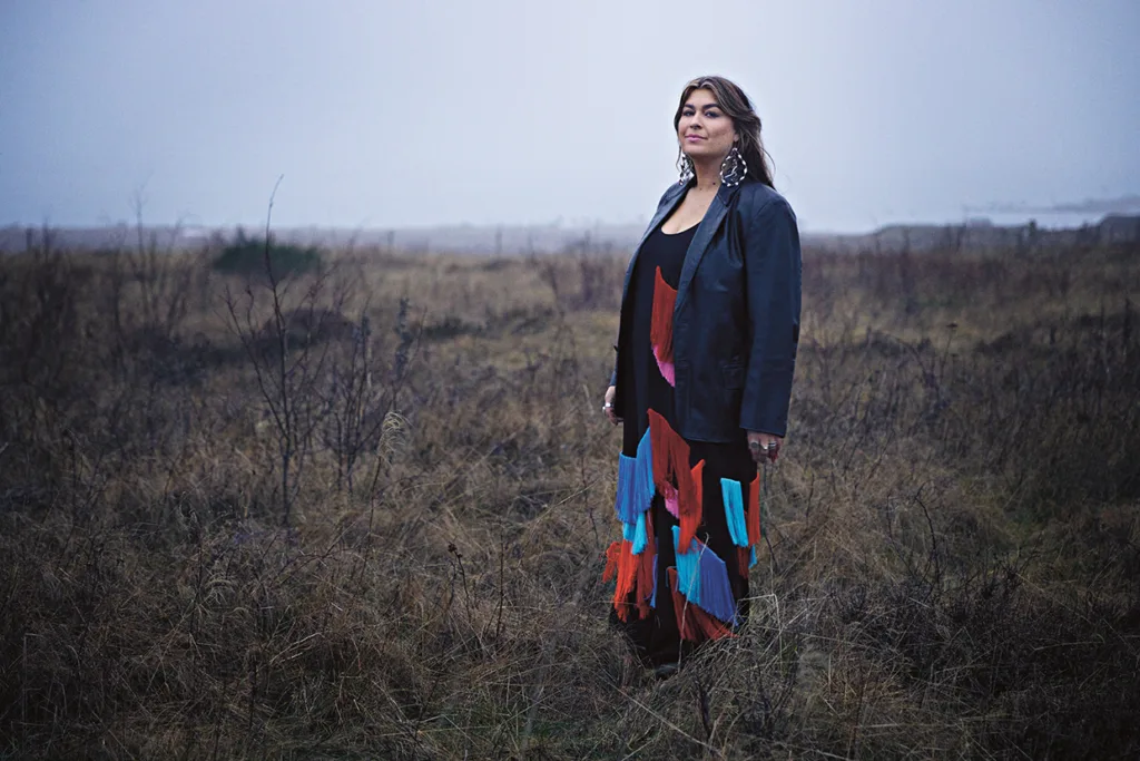 Linnea Henriksson står på ett gräsbevuxet, dimmigt fält iklädd en färgglad klänning med fransar och en svart skinnjacka. Hon ser självsäkert framåt, med stora örhängen. Bakgrunden är dimmig och mulen, vilket skapar en lugn och stämningsfull atmosfär.