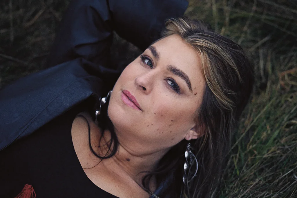 Linnea Henriksson med långt brunt hår och ljusa slingor ligger på en gräsmatta och tittar in i kameran. Hon bär en mörk jacka, stora silverörhängen och naturlig makeup, med armen vilande bakom huvudet.