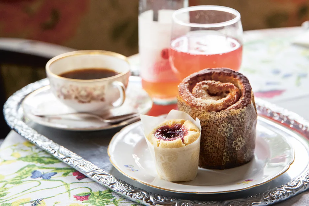 På en silverbricka står en kopp kaffe, ett glas rosa drink, en rulltårta pudrad med socker och en liten muffins toppad med röd sylt, allt på en blommönstrad duk.