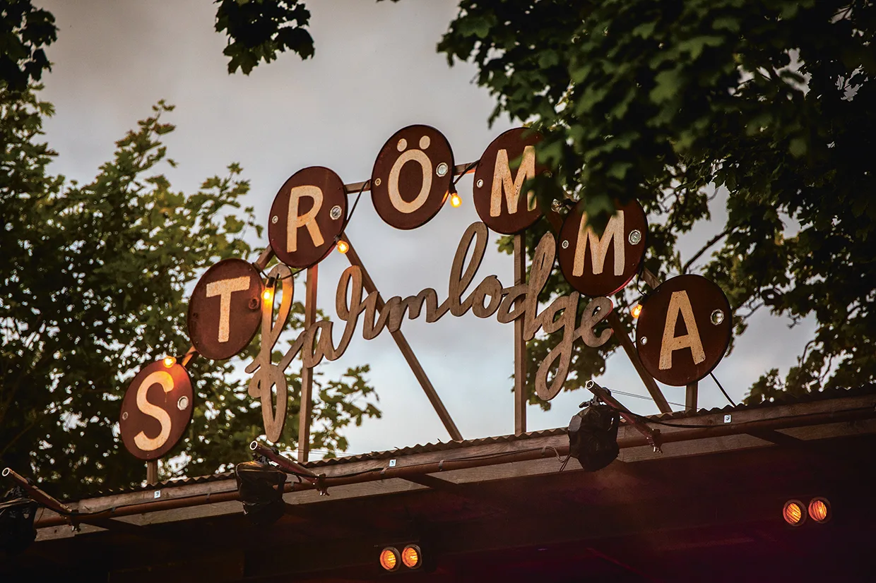 En skylt med texten "Strömma Farmlodge" i dekorativa bokstäver lyses upp av små glödlampor, omgiven av gröna träd och mot en molnig himmel.