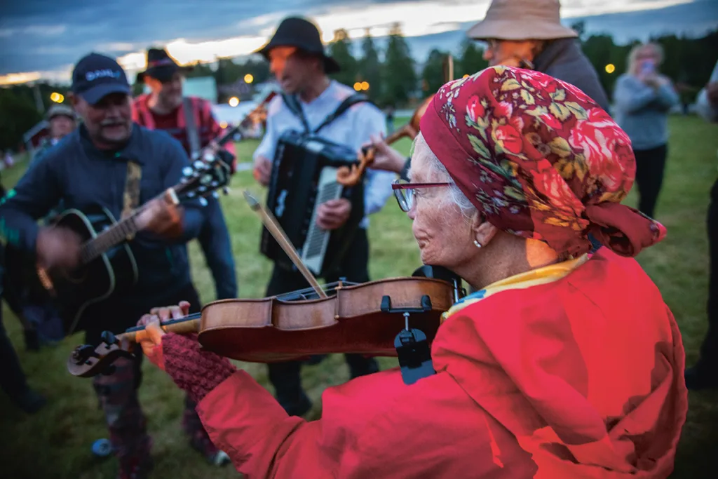 En grupp människor spelar instrument utomhus i skymningen, bland annat en äldre kvinna med röd huvudduk och glasögon som spelar fiol, med andra på gitarr och dragspel, omgiven av gräs och träd.