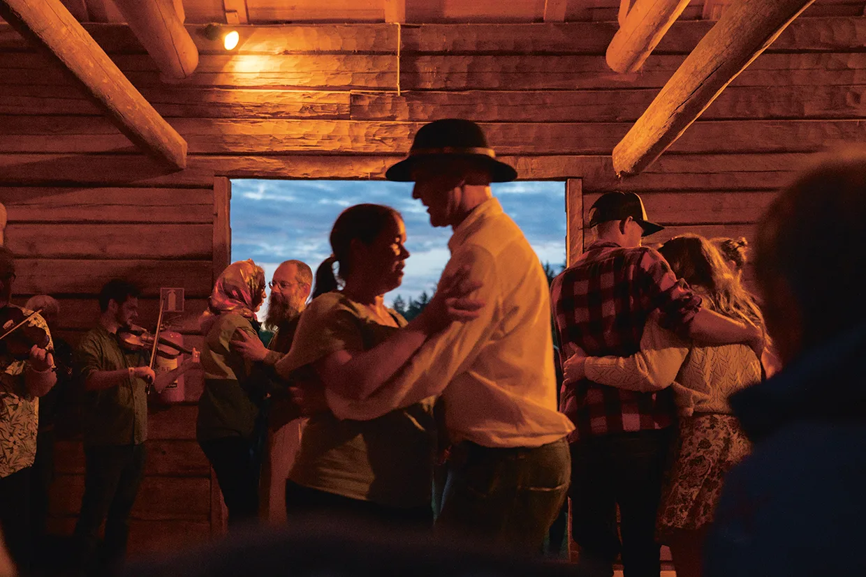 Människor dansar i par i en rustik lada i skymningen, upplyst av varmt inomhusljus. Ett fönster visar kvällshimlen och en musiker spelar violin vid sidan om.