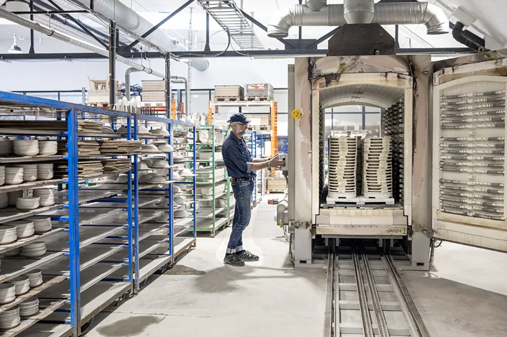 En arbetare i en keramikfabrik står bredvid en industriell ugn med keramikplattor på ställningar; hyllor fyllda med fler plattor finns på båda sidor av den väl upplysta arbetsplatsen.