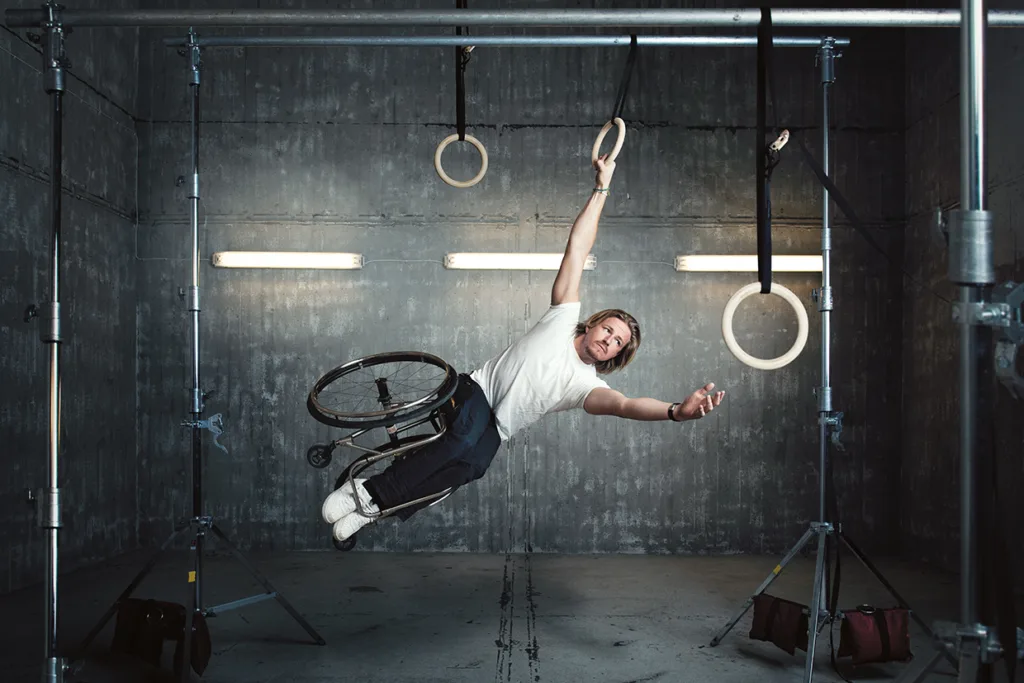 En man i rullstol hänger med en arm i gymnastikringar i ett gym i industriell stil, balanserar i luften och ler självsäkert.