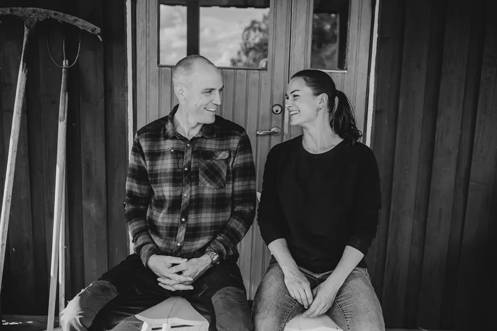 Björn Ferry & Heidi Andersson sitter sida vid sida på en veranda och ler mot varandra. Båda bär vardagskläder; mannen har en rutig skjorta och kvinnan en mörk tröja. Trädgårdsredskap står lutade mot träväggen bakom dem.