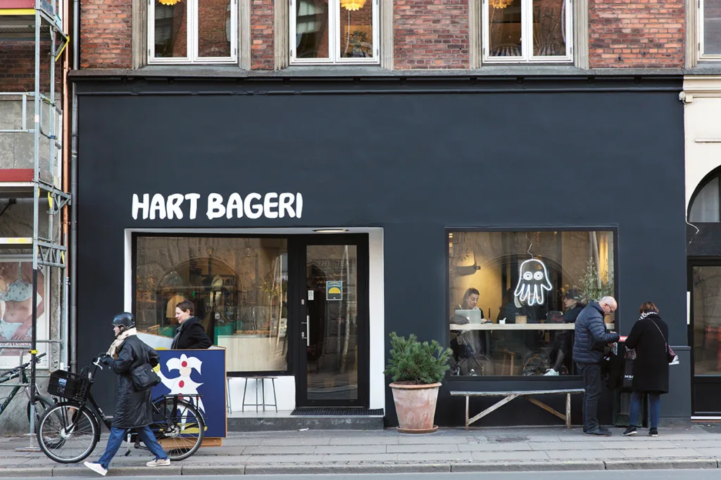 Ett modernt bageri med en svart fasad med texten HART BAGERI. Människor står utanför, vissa med cyklar, medan andra sitter inne vid ett stort fönster med en vit spökliknande teckning.