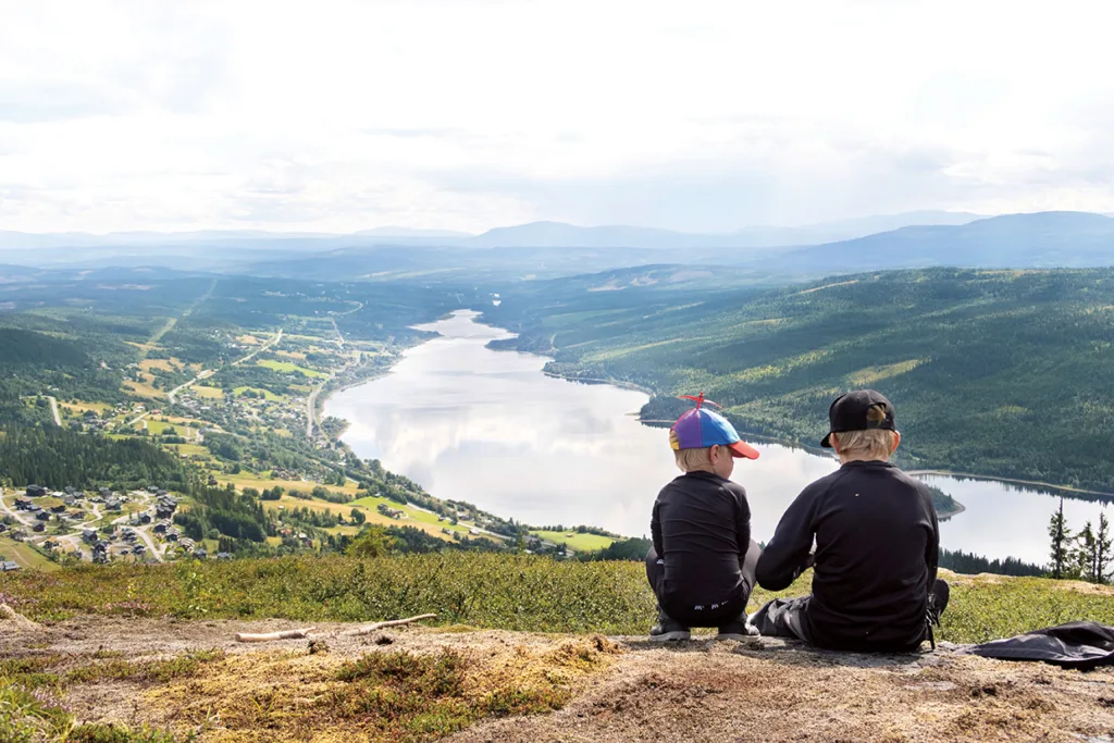 Två barn sitter på en kulle med utsikt över en slingrande flod, ett skogslandskap och avlägsna berg under en molnig himmel. Hus och vägar syns i dalen nedanför.