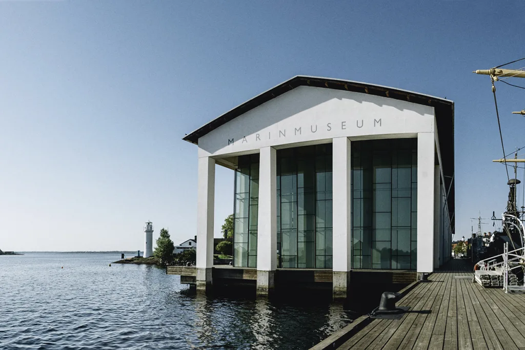 En modern vit byggnad med beteckningen MARINMUSEUM står vid vattnet, delvis över vattnet, med stora glasfönster och en klarblå himmel ovanför. En fyr och grönska syns i bakgrunden.