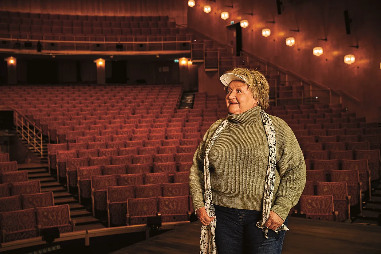 En kvinna i grön tröja, halsduk och keps står på en scen och ler, med tomma röda teatersäten och varma vägglampor synliga i bakgrunden.