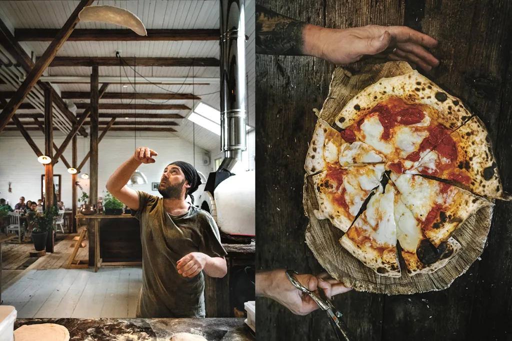 En man kastar pizzadeg i luften inne på en rustik pizzeria; bredvid honom håller en hand en vedeldad pizza med smält ost, pepperoni och förkolnad skorpa, placerad på en serveringsbräda av trä.