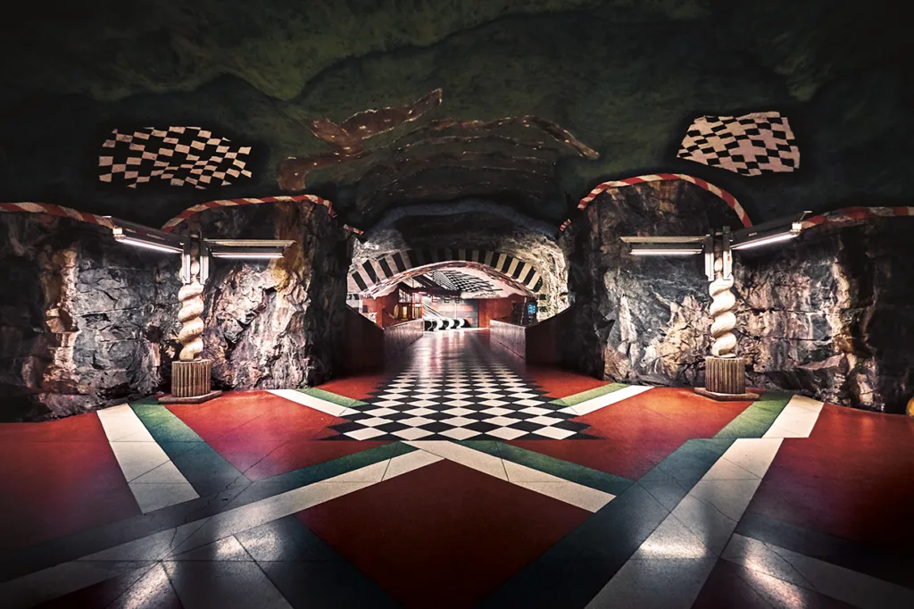 En underjordisk tunnelbanestation med ett grottliknande tak, exponerade bergväggar och ett geometriskt svart, vitt och rött mönstrat golv. Stationen har djärva, färgglada mönster och upplysta pelare.