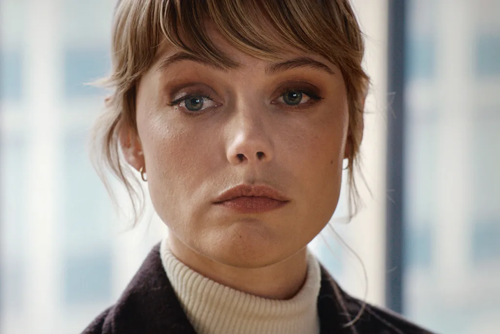 Frida Gustavsson med kort blont hår och lugg tittar rakt in i kameran med ett neutralt uttryck. Hon är klädd i en krämfärgad polotröja och mörk kavaj, med mjukt upplysta fönster i bakgrunden.