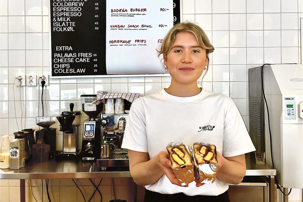 En kvinna i vit skjorta står på ett café, ler och håller i två smörgåsar. Bakom henne finns en kaffemaskin, olika tillbehör och en menytavla mot en vit kaklad vägg.