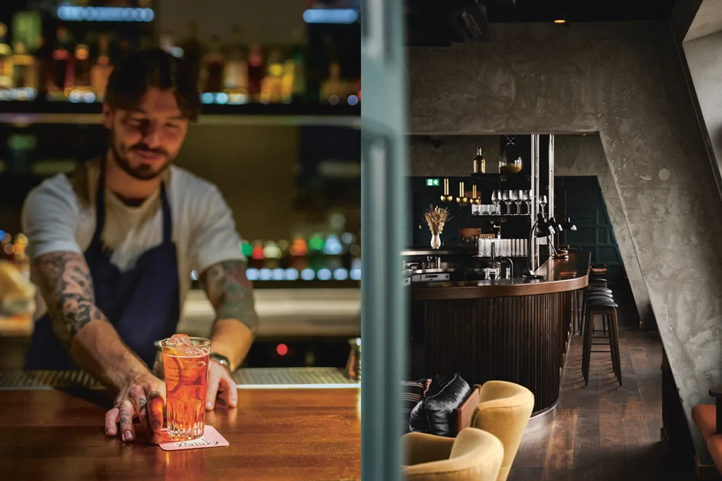En bartender med tatueringar och förkläde serverar en cocktail i en bar; bredvid honom syns en svagt upplyst, stilfull barinredning med trämöbler och modern dekor.