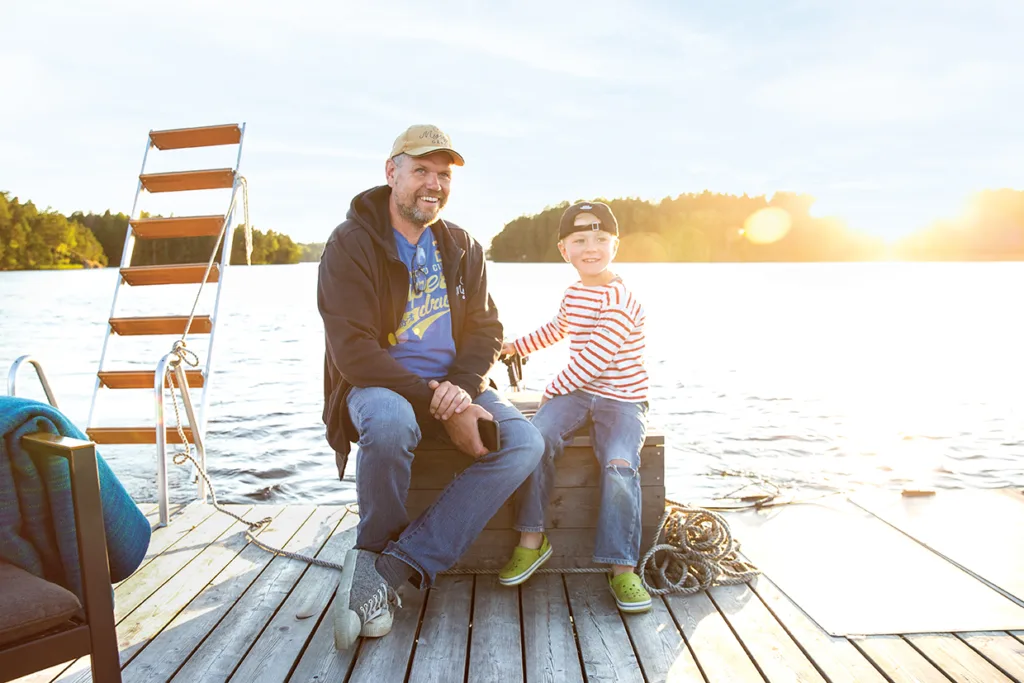 En leende man och ett barn sitter på en träbrygga vid en sjö och njuter av solen. Mannen bär keps och huvtröja, medan barnet bär randig skjorta och bakvänd keps. Vatten och träd syns i bakgrunden.