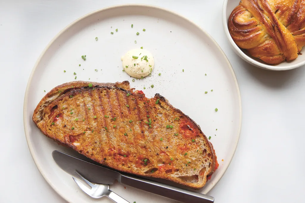 En grillad brödskiva garnerad med örter ligger på en vit tallrik och serveras med en klick krämig sås. Besticken vilar på tallriken. I en skål bredvid ligger ett snurrat bakverk.