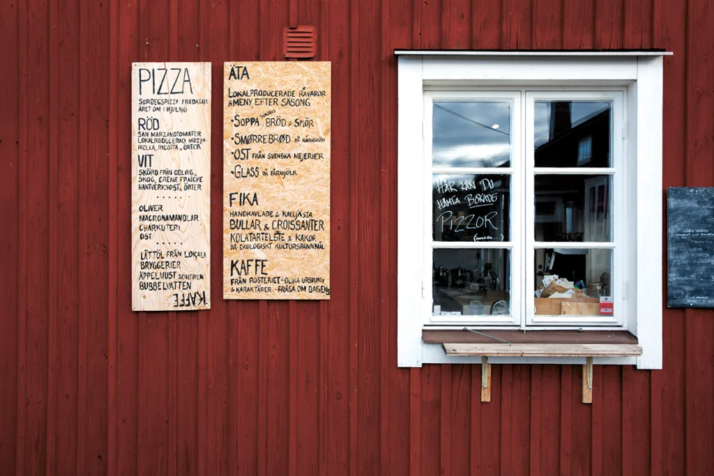 En röd träbyggnad med två menytavlor som listar pizzor och andra maträtter på svenska, bredvid ett vitkarmat fönster med en liten öppen disk under. Några köksartiklar syns inuti genom fönstret.
