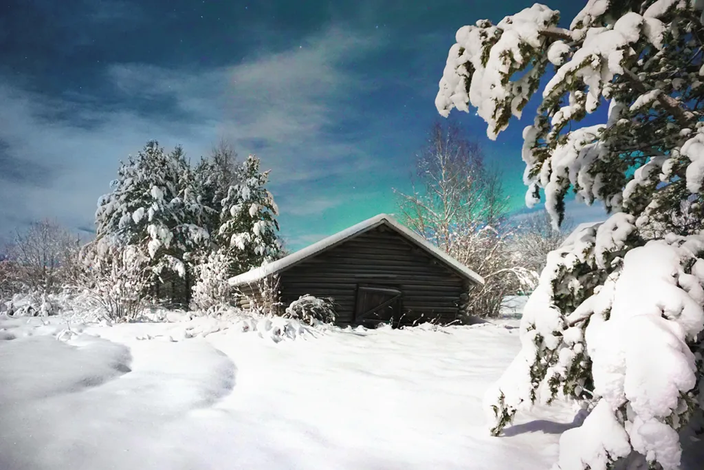 En liten trästuga ligger i ett snöigt landskap, omgiven av snötäckta träd under en stjärnklar himmel med en antydan till grönt norrsken. Snön täcker marken och skapar en fridfull vinterscen.