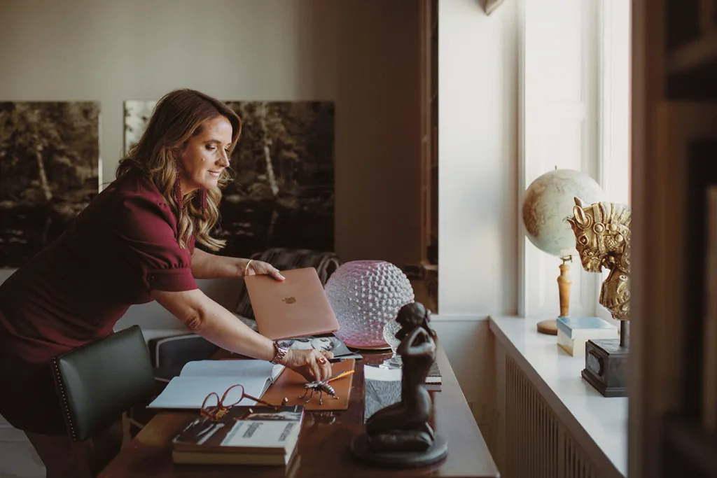 Denise Rudberg i vinröd klänning står vid ett skrivbord och lägger ner en bärbar dator. På skrivbordet finns böcker, glasögon och statyetter. Solljuset flödar in genom ett fönster med en jordglob och prydnadsföremål på fönsterbrädan.