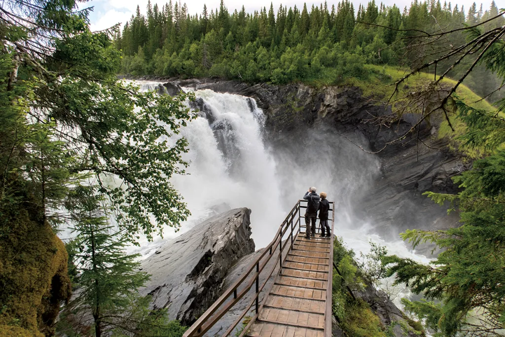 Två personer står på en träbrygga med utsikt över ett stort, mäktigt vattenfall omgivet av grönskande träd och klippor. Tät skog fyller bakgrunden under en delvis molnig himmel.