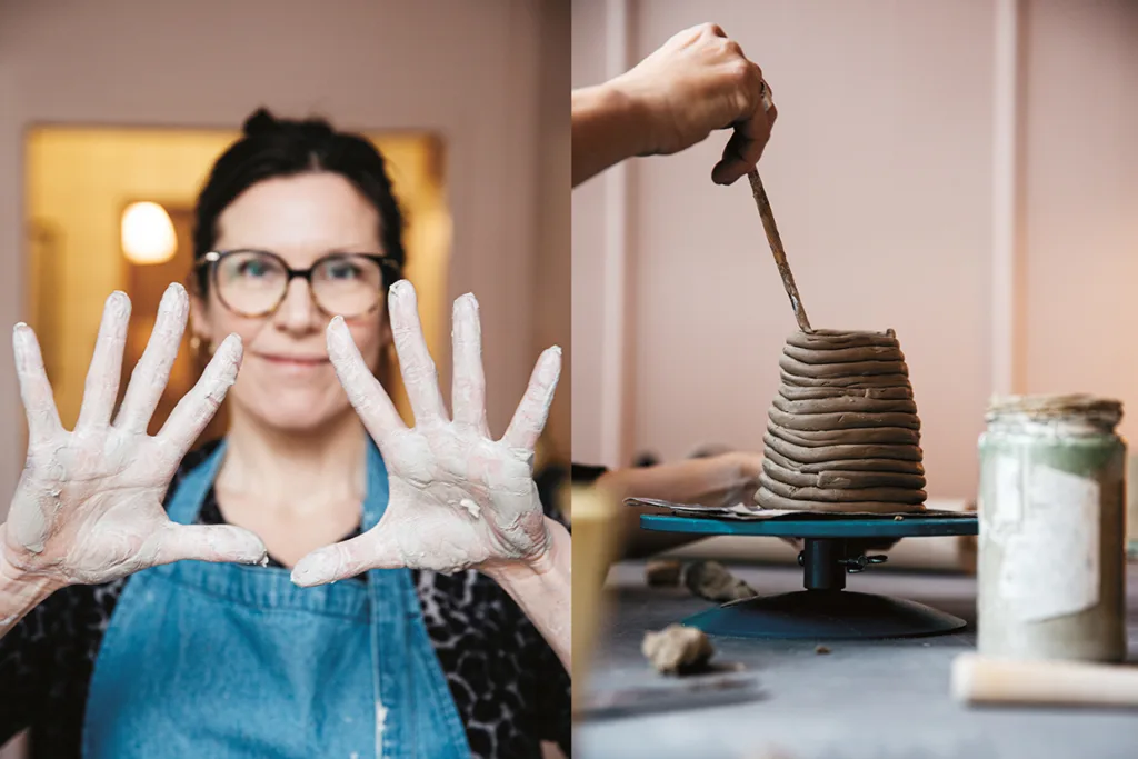 En kvinna i glasögon och förkläde håller upp sina lertäckta händer; bredvid henne formar händerna en upprullad lerkruka på en drejskiva i en keramikstudio.