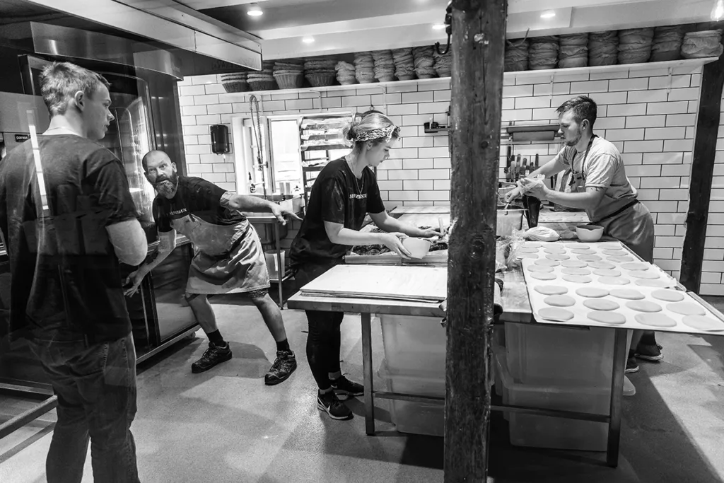 Fyra personer arbetar i ett hektiskt kök med att tillverka pizzadeg. En person står till vänster, två arbetar vid ett förberedelsebord med att rulla deg och Johan Sörberg gestikulerar livligt. I bakgrunden syns högar med deg och köksutrustning.