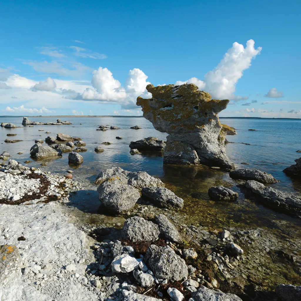 Klippig strandlinje med stora, unikt formade havstaplar av kalksten (rauks) som reser sig ur grunt, klart vatten under en blå himmel med spridda moln.
