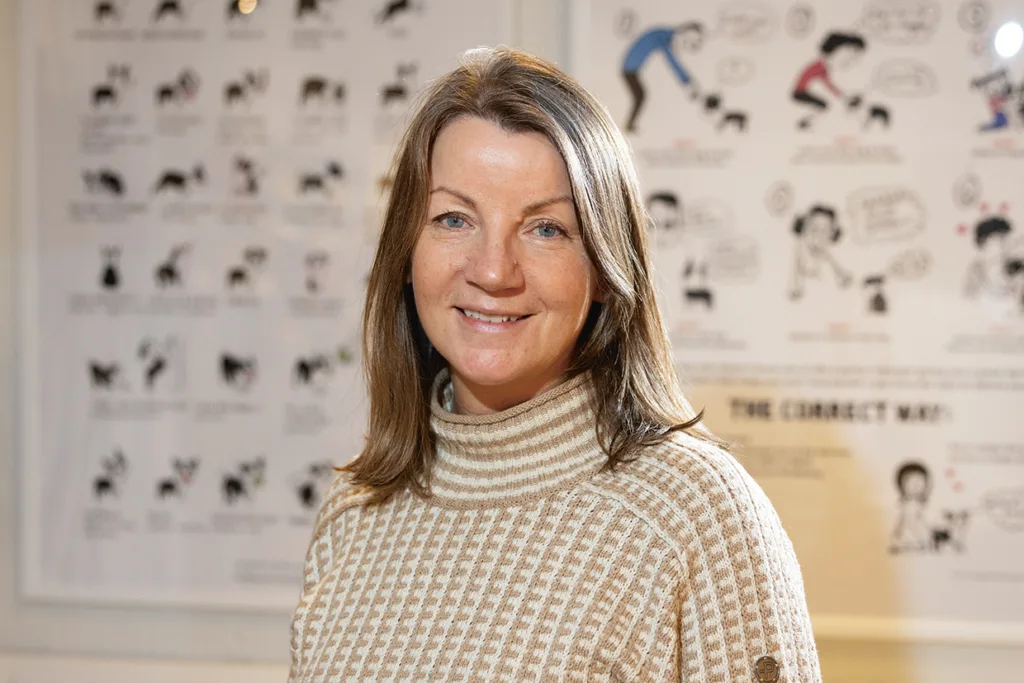 Kicki Fellstenius med ljusbrunt hår i beige rutig polotröja ler mot kameran, stående inomhus med illustrerade affischer suddiga i bakgrunden.