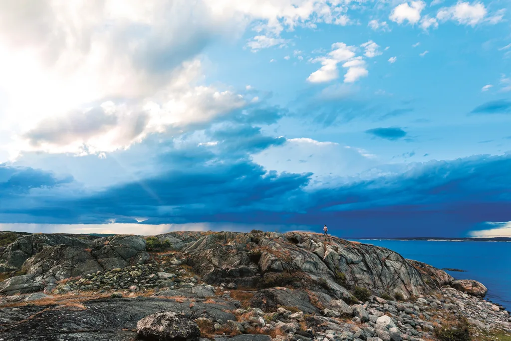 Klippig kust under en dramatisk himmel med solstrålar som skiner genom molnen; en person står på klipporna och blickar ut över det blå havet en klar, delvis molnig dag.