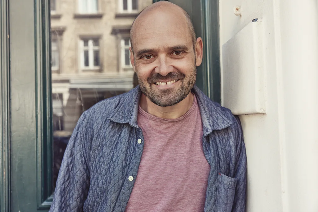En leende, skallig David Dencik med trimmat skägg i rosa T-shirt och blå quiltad skjorta står utomhus mot en vit vägg med en grön dörr och byggnadsreflexer i bakgrunden.