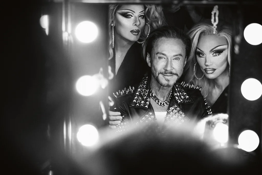 Svartvitt foto av tre personer, två i dramatisk dragmakeup och peruker, och Christer Lindarv med skägg och spikjacka, som poserar tillsammans och ler framför en upplyst sminkspegel.